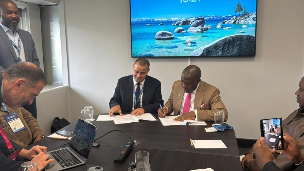 An indoor, eye-level shot of a formal signing ceremony inside a modern conference room. Two men sit at a dark wooden table signing white documents. The man on the left wears a dark suit and tie, while the man on the right wears a beige suit jacket, a bright pink tie, and a white collared shirt. A large flat-screen monitor mounted on the wall behind them displays a scenic tropical beach background under a blue sky. Other attendees are present, including a man on the far left looking down at a laptop computer and another on the far right holding up a black smartphone to take a photo of the signing.