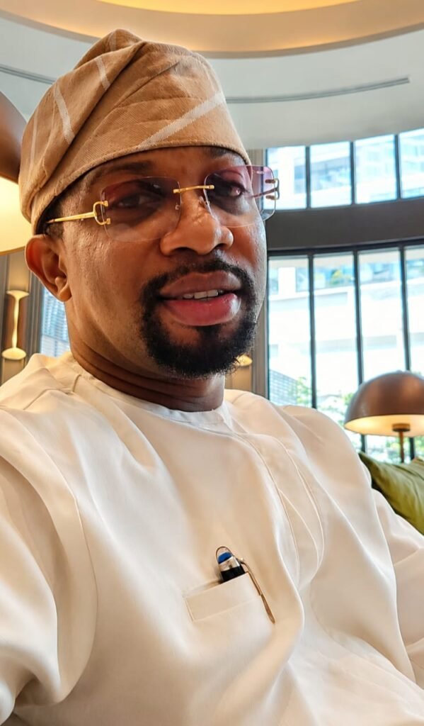 A close-up portrait of David Adesanya, Group Managing Director of Elite Logistics & Development Services Limited, wearing a traditional light-colored Nigerian agbada and a matching cap, with gold-rimmed glasses.