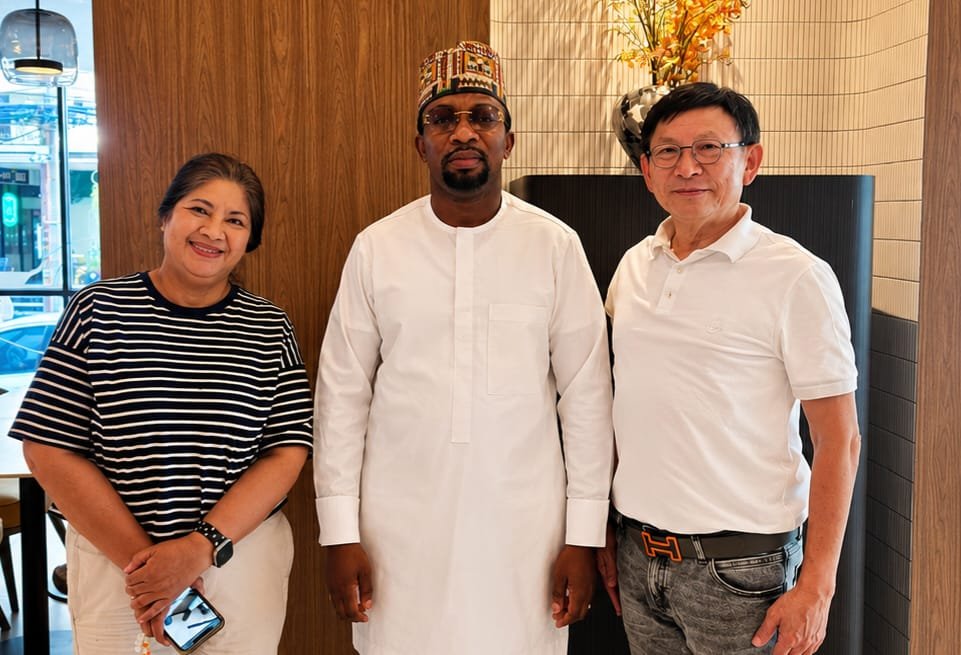 David Adesanya standing in the center in a white native outfit and embroidered cap, flanked by two international business partners (a woman in a striped shirt on the left and a man in a white polo shirt on the right) in a modern indoor setting.